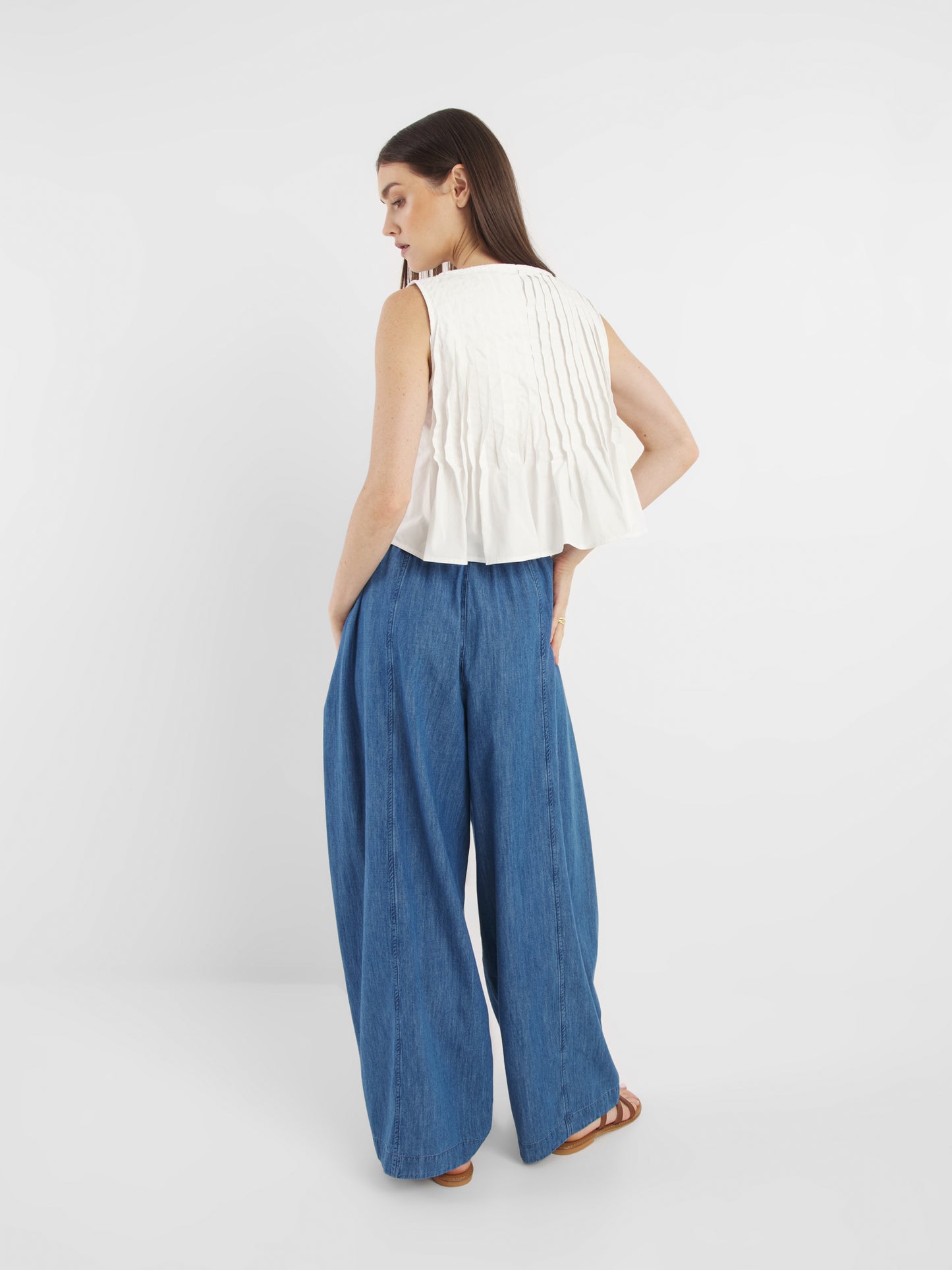 Eine Frau mit langen dunklen Haaren trägt ein ärmelloses, plissiertes weißes Oberteil und eine weit geschnittene Lizzy-Denim-Hose, dazu braune Sandalen. Sie steht mit dem Gesicht zur Seite vor einem schlichten weißen Hintergrund.