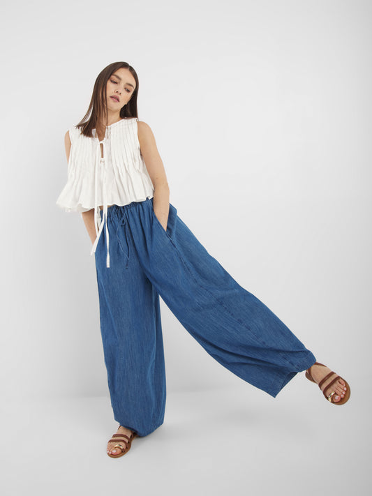 Eine Frau modelliert die blaue Lizzy Denim-Hose mit weitem Bein, ein ärmelloses weißes Oberteil und braune Sandalen. Sie steht mit einem angewinkelten Bein und den Händen in den Taschen vor einem schlichten, hellen Hintergrund.