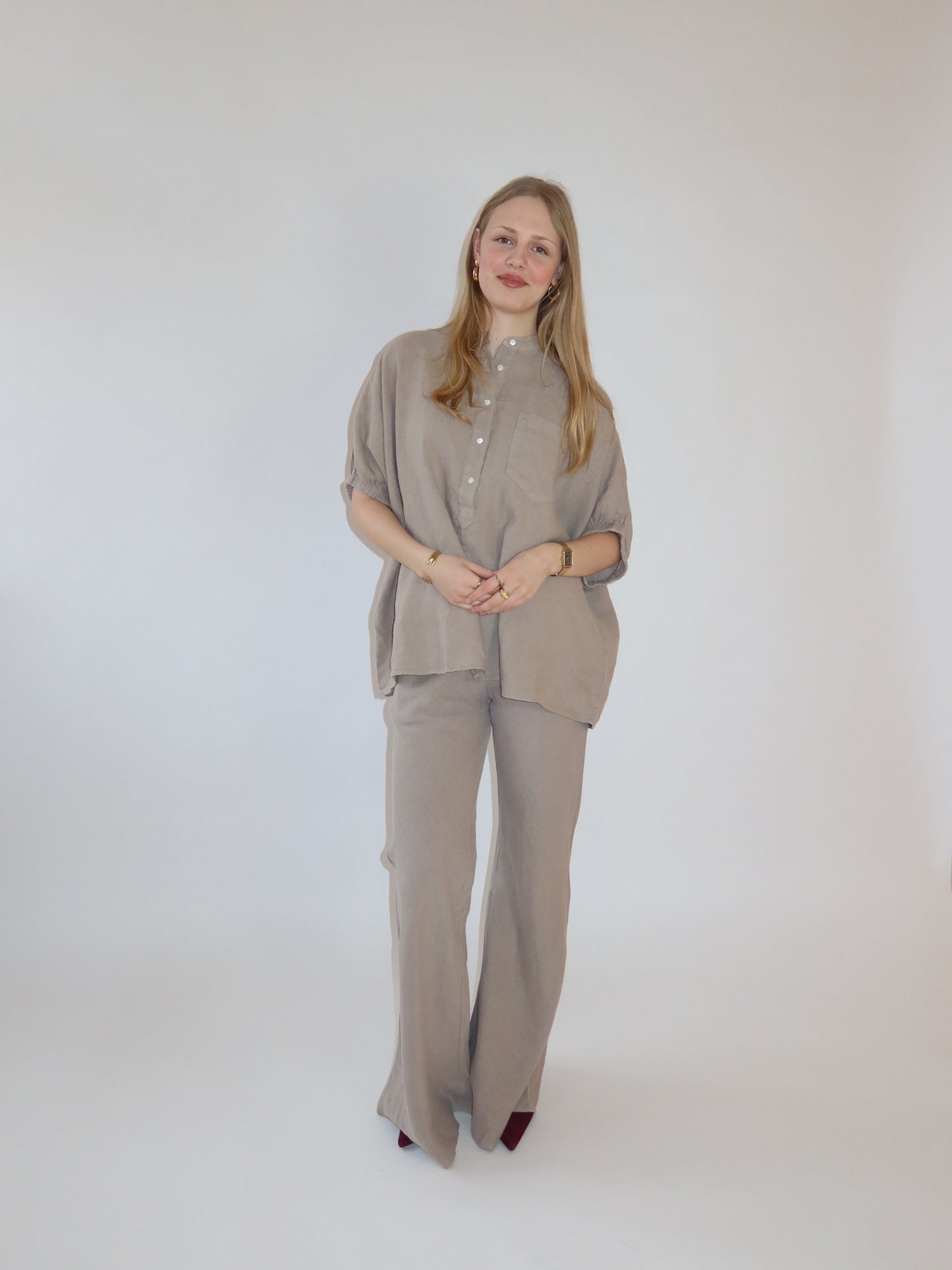 Eine Frau mit langen blonden Haaren steht vor einem weißen Hintergrund und trägt das Bluse Cowboy Button Leinenhemd mit passender Hose mit weitem Bein. Sie lächelt sanft, die Hände vor sich verschränkt.