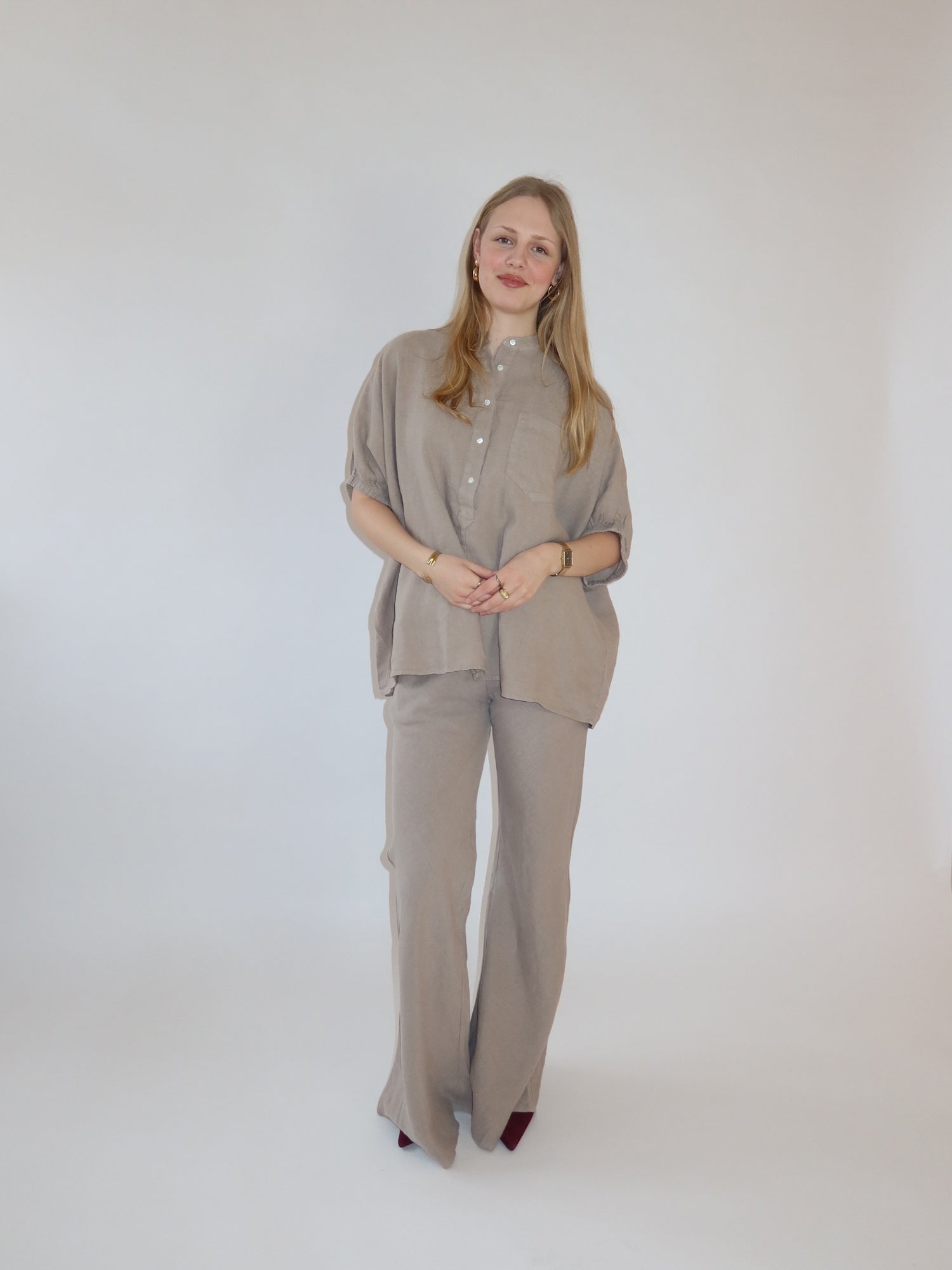 Eine Frau mit langen blonden Haaren steht vor einem weißen Hintergrund und trägt das Bluse Cowboy Button Leinenhemd mit passender Hose mit weitem Bein. Sie lächelt sanft, die Hände vor sich verschränkt.