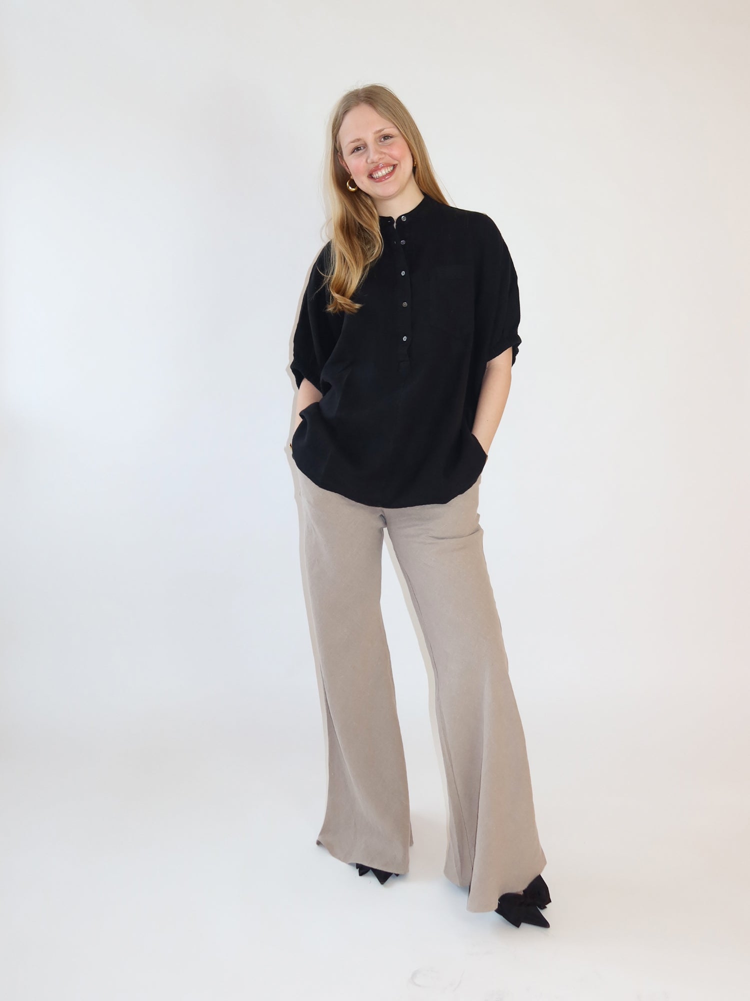 Eine Frau mit langen blonden Haaren lächelt vor einem weißen Hintergrund und trägt die Bluse Cowboy Button Linen Black und tan Wide-Leg Pants-perfekt als Leinenbluse Damen für den Sommer. Ihre Hände stecken in den Taschen.