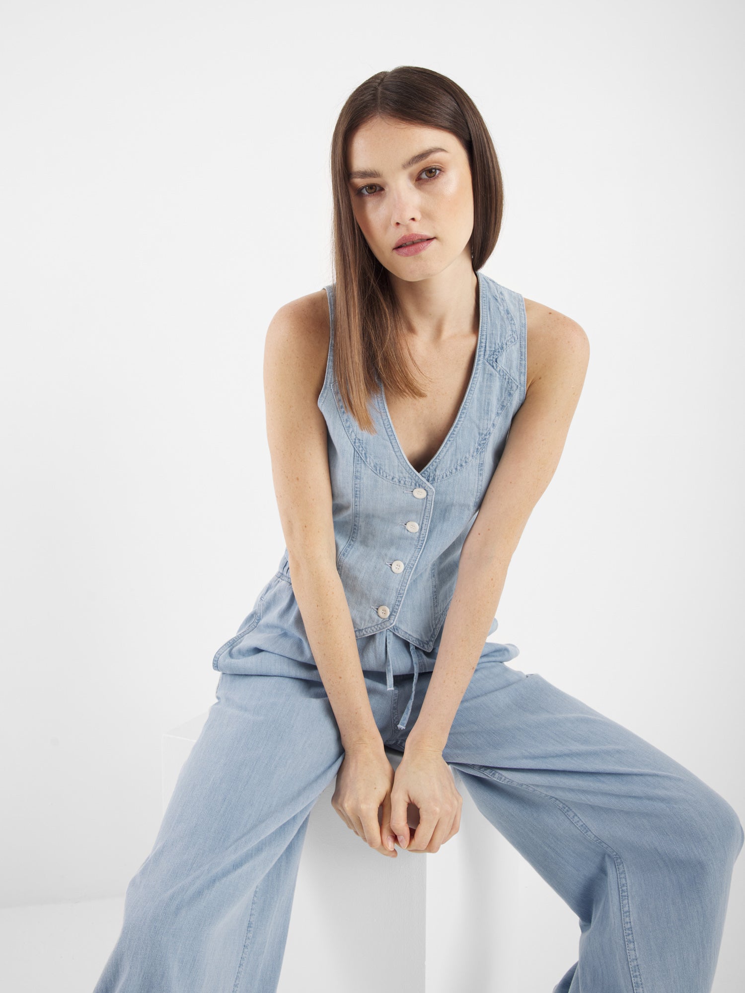 Eine Frau mit glattem braunem Haar sitzt auf einem weißen Block und trägt die Vest Denim - ein ärmelloses hellblaues Jeansoberteil mit Knöpfen und einer dazu passenden Hose mit weitem Bein - und schaut mit neutralem Blick direkt in die Kamera.