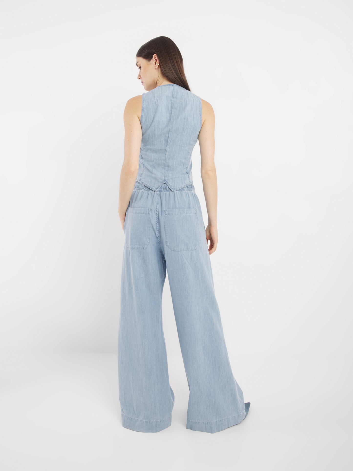 Eine Frau mit langem, glattem, dunklem Haar steht vor einem schlichten weißen Hintergrund und trägt die ärmellose, helle Jeansweste Vest Denim und die dazu passende Jeans mit weitem Bein.