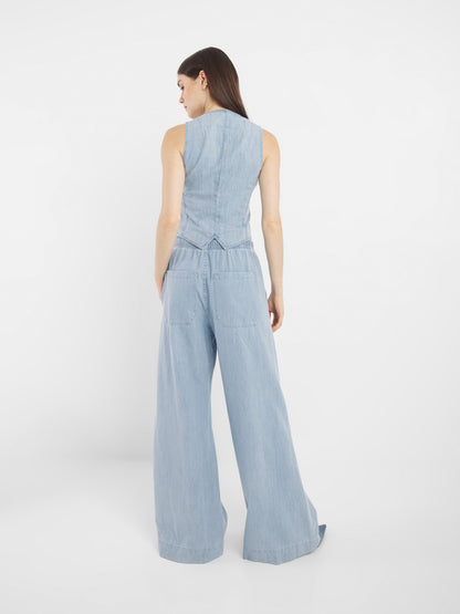 Eine Frau mit langem, glattem, dunklem Haar steht vor einem schlichten weißen Hintergrund und trägt die ärmellose, helle Jeansweste Vest Denim und die dazu passende Jeans mit weitem Bein.