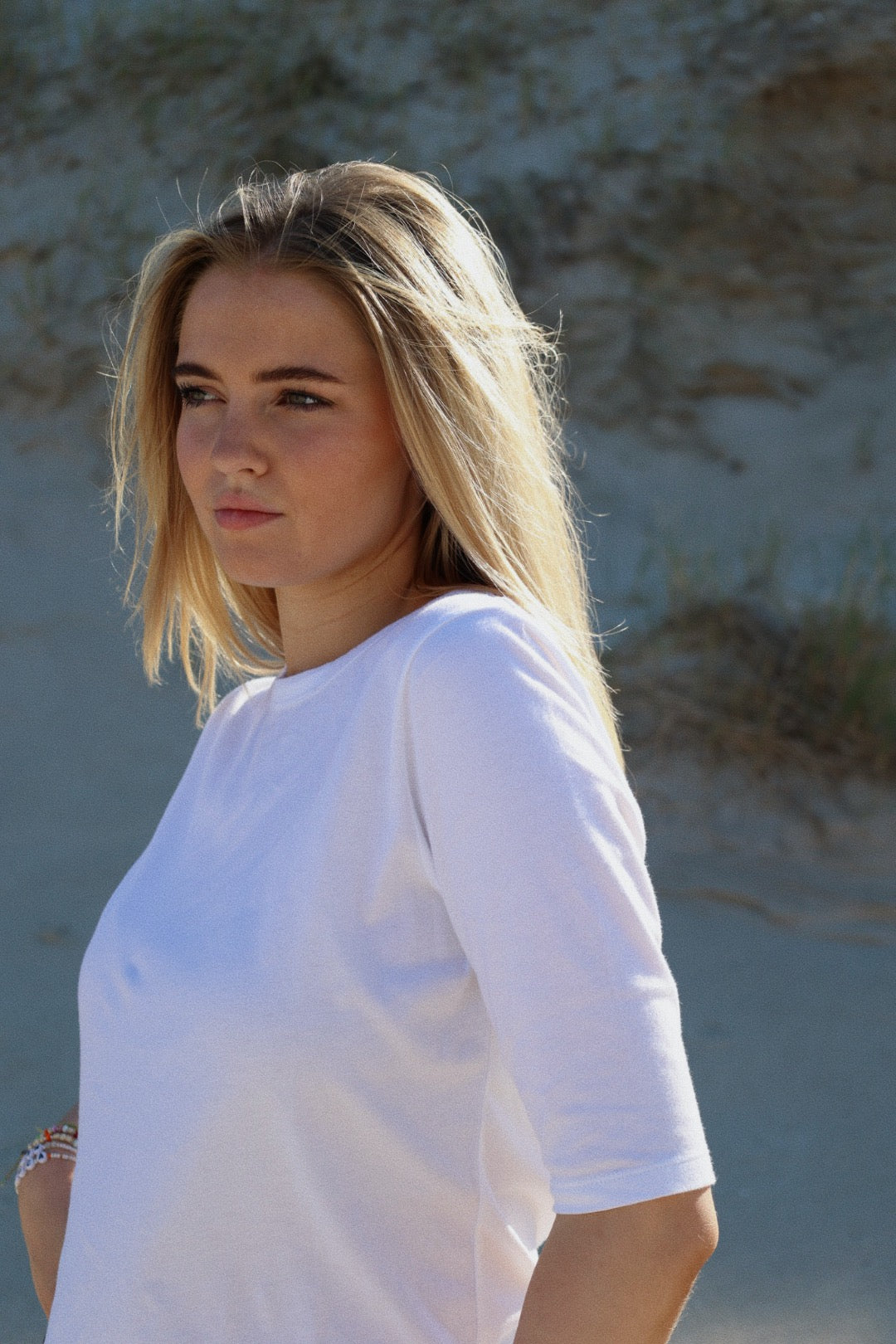 Eine junge Frau mit langen blonden Haaren trägt ein T-Shirt Half Sleeve White aus 100 % Baumwolle. Sie steht im Freien im Sonnenlicht mit Sand und spärlichem Gras im Rücken und blickt nachdenklich in die Ferne.