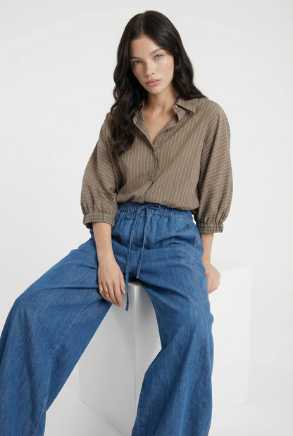 Eine Frau mit langen dunklen Haaren sitzt auf einem weißen Block, trägt das Antonia Short Sleeve Taupe Stripes Shirt und eine blaue Jeanshose mit weitem Bein und schaut mit neutralem Blick direkt in die Kamera.