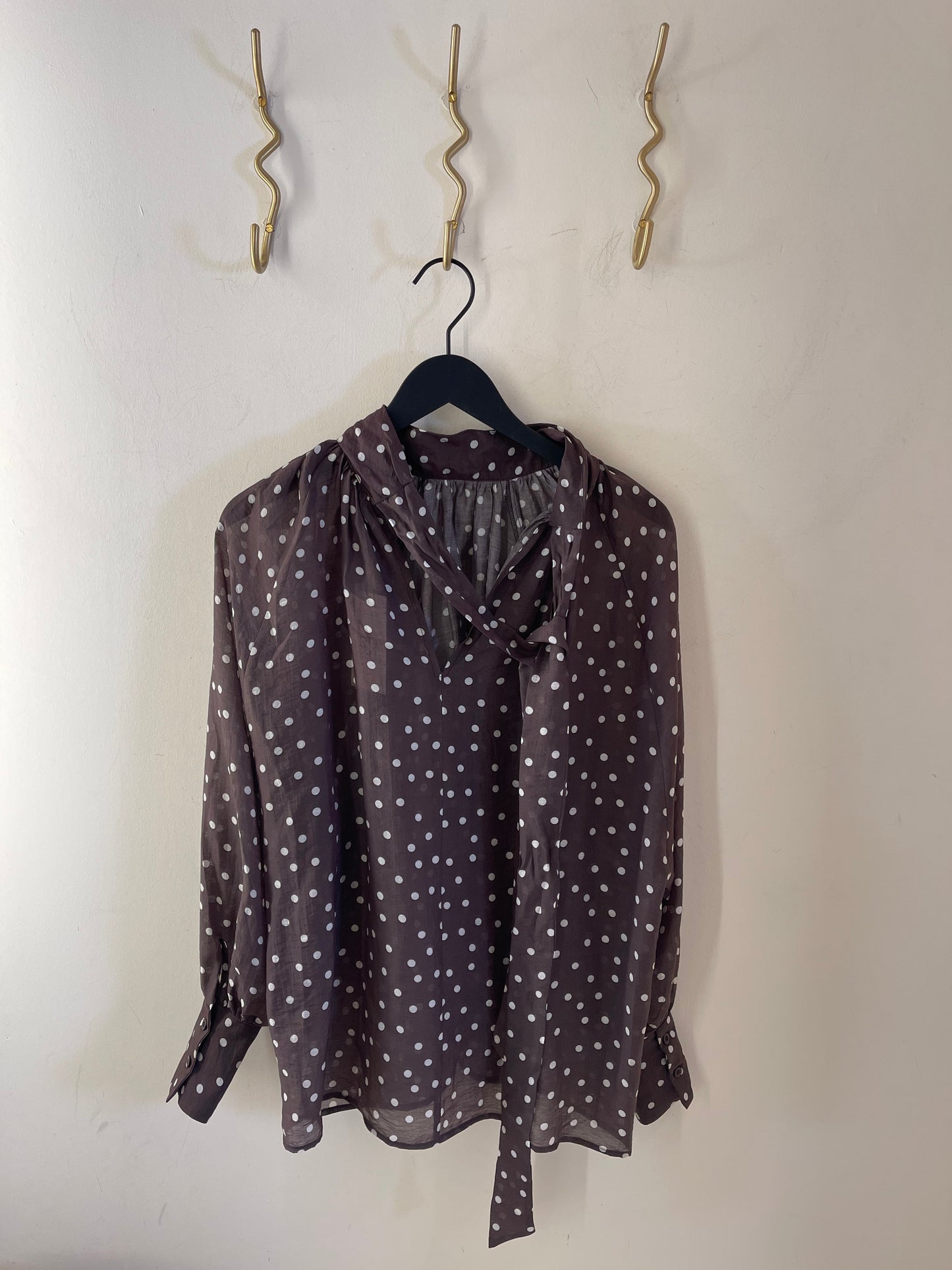 Die Bluse „Dotty Bow Grey Latte“, eine schicke, in Italien gefertigte Langarmbluse mit Polka-Dots aus weichem Baumwoll-Voile, hängt an einem schwarzen Kleiderbügel vor einer weißen Wand. Weiße Punkte zieren den grau-lattefarbenen Stoff, während darüber drei goldene, geschwungene Haken angebracht sind.