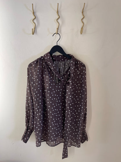 Die Bluse „Dotty Bow Grey Latte“, eine schicke, in Italien gefertigte Langarmbluse mit Polka-Dots aus weichem Baumwoll-Voile, hängt an einem schwarzen Kleiderbügel vor einer weißen Wand. Weiße Punkte zieren den grau-lattefarbenen Stoff, während darüber drei goldene, geschwungene Haken angebracht sind.