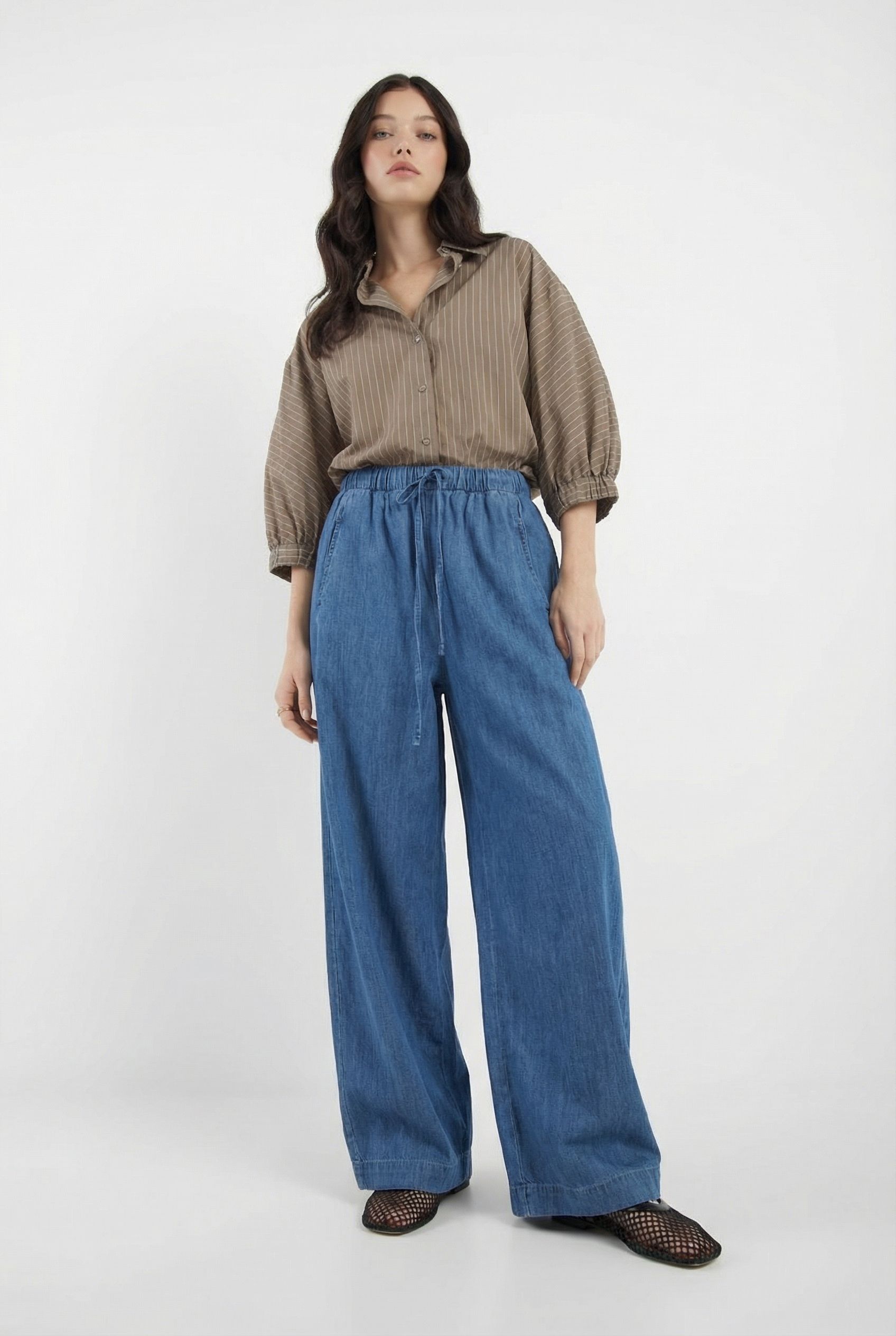 Eine Frau mit langen, gewellten dunklen Haaren und einem neutralen Gesichtsausdruck trägt das Antonia Short Sleeve Taupe Stripes Shirt mit weit geschnittenen Blue Jeans und schwarzen Webschuhen vor einem schlichten weißen Hintergrund.