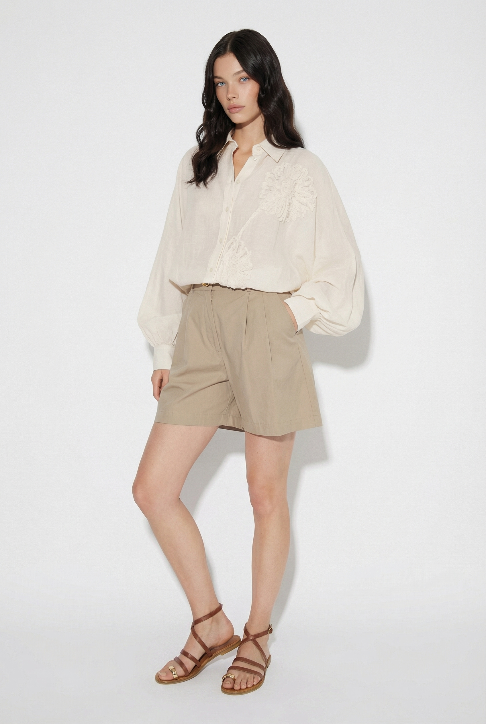 Eine Frau mit langen dunklen Haaren posiert vor einem weißen Hintergrund und trägt die Bluse Antonia Crafted Panna, eine lockere cremefarbene Bluse mit Blumendetails, gepaart mit hellbraunen High-Waisted-Shorts und braunen Riemchensandalen.