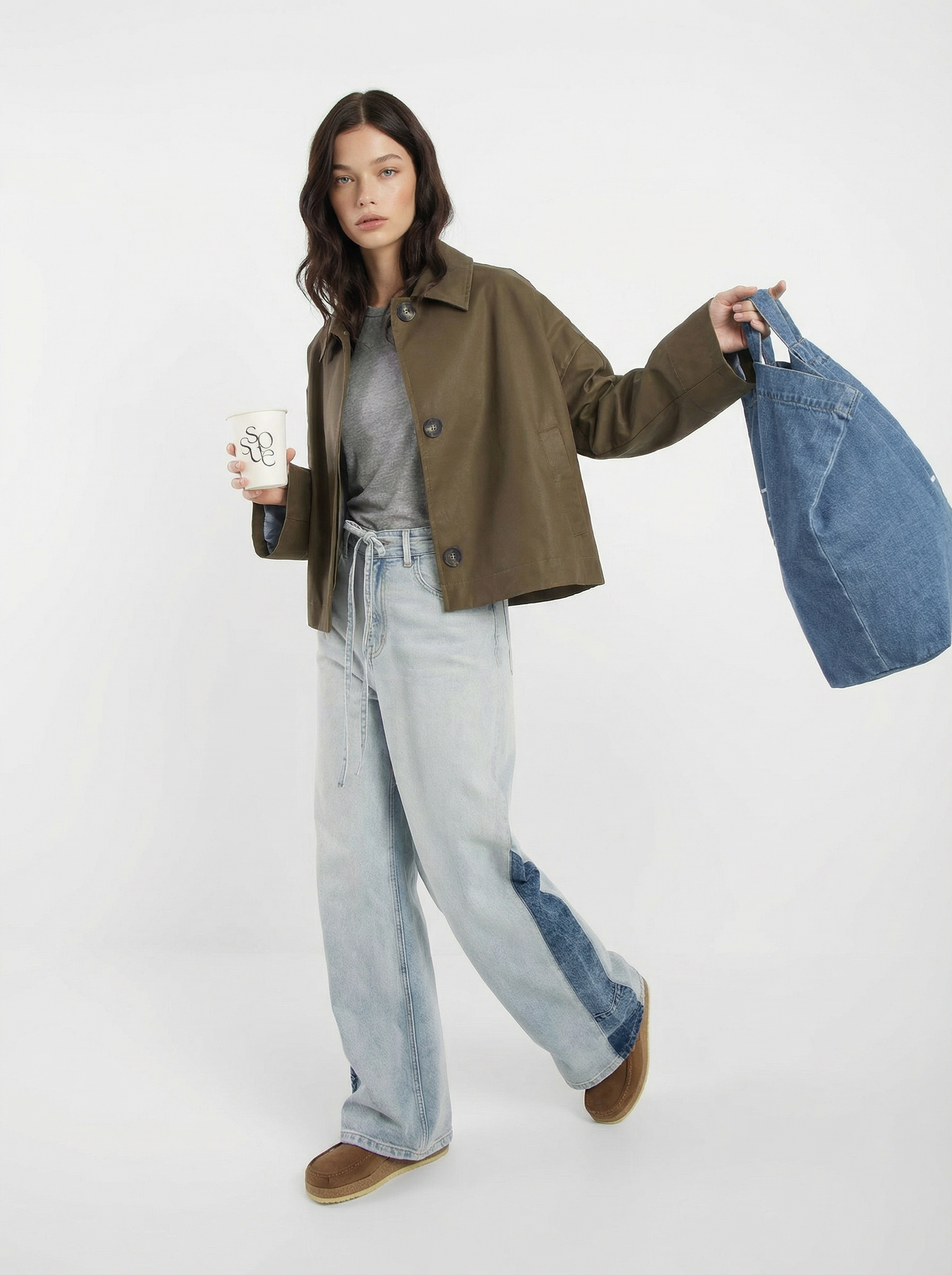 Eine Frau in einem grünen Dog Walker Coat, einem grauen Hemd und hellblauen Jeans mit weitem Bein hält eine Kaffeetasse und eine blaue Denim-Tasche, während sie vor einem schlichten weißen Hintergrund steht.