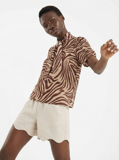 Eine Person trägt die Bluse Bowy Slim Zebra Brown, eine braune und beigefarbene kurzärmelige Bluse mit Zebradruck und einer Krawatte, gepaart mit hellbeigen Shorts mit Wellenmuster, und posiert vor einem weißen Hintergrund.