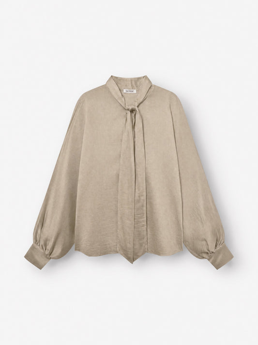 Die Bluse „Antonia Bow Taupe“ besticht durch ihren beigen Farbton, lange Ärmel, eine lockere Passform, einen Bindegürtel am Halsausschnitt, geknöpfte Ärmelbündchen und einen dezenten Schimmer – hier auf weißem Hintergrund abgebildet.