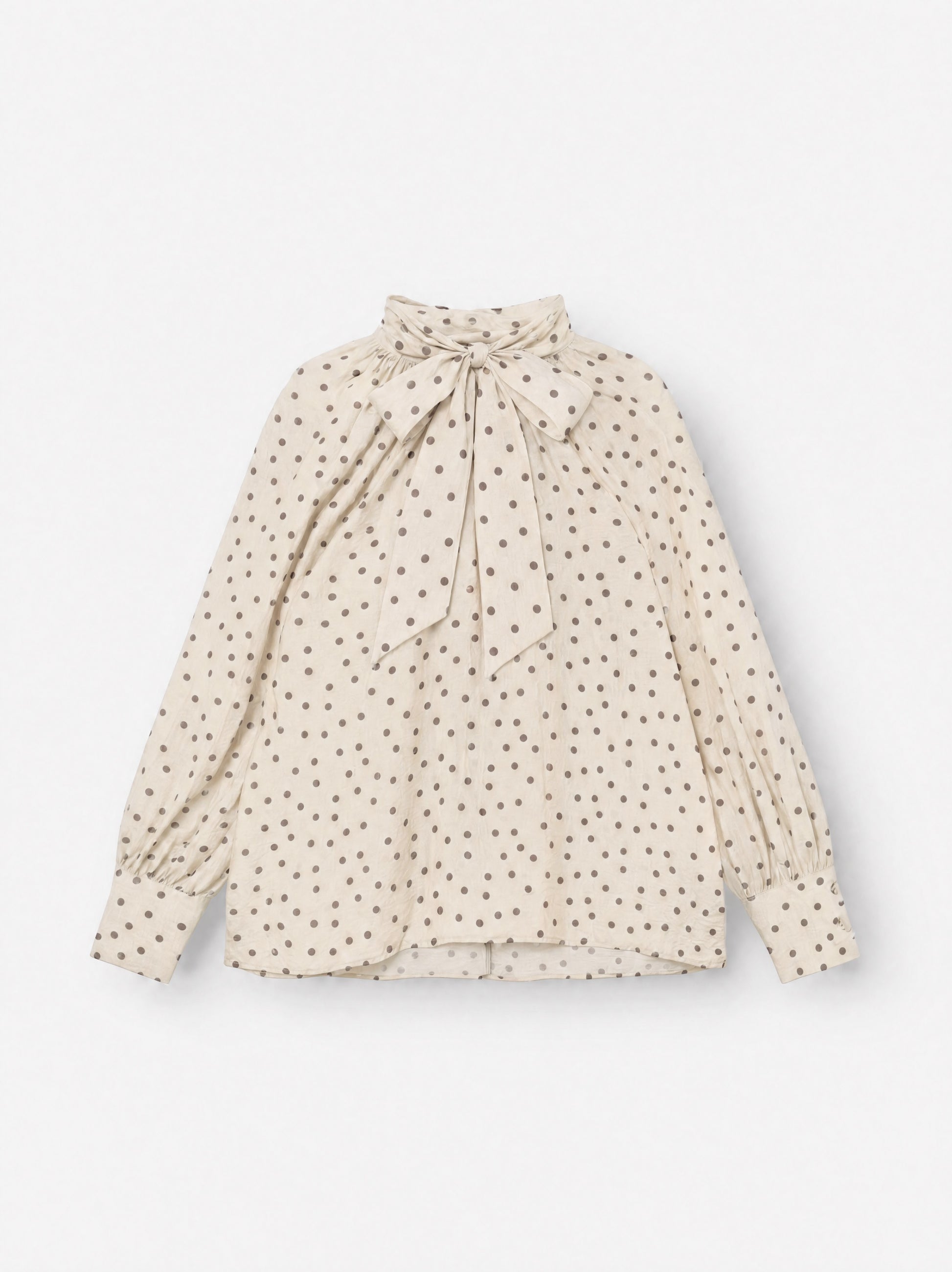 Die Bluse „Dotty Bow Ecru Brown“ ist eine langärmelige Bluse mit braunen Polka-Punkten, einem hohen Ausschnitt und einer übergroßen Schleife, die am Hals gebunden wird. Sie wurde in Italien aus feinem Baumwoll-Voile gefertigt und verfügt über geknöpfte Ärmelbündchen sowie eine lockere, fließende Passform.