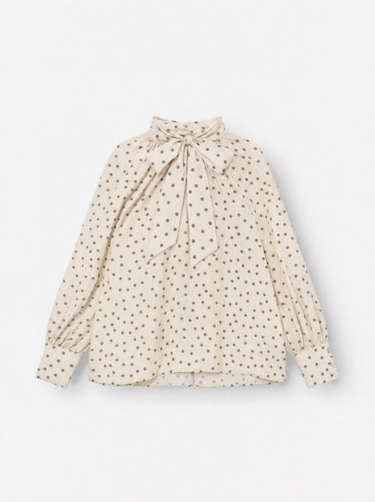 Die Bluse „Dotty Bow Ecru Brown“ ist eine langärmelige Bluse mit braunen Polka-Punkten, einem hohen Ausschnitt und einer übergroßen Schleife, die am Hals gebunden wird. Sie wurde in Italien aus feinem Baumwoll-Voile gefertigt und verfügt über geknöpfte Ärmelbündchen sowie eine lockere, fließende Passform.