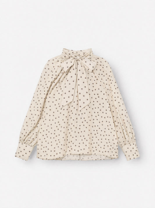 Die Bluse „Dotty Bow Ecru Brown“ ist eine langärmelige Bluse mit braunen Polka-Punkten, einem hohen Ausschnitt und einer übergroßen Schleife, die am Hals gebunden wird. Sie wurde in Italien aus feinem Baumwoll-Voile gefertigt und verfügt über geknöpfte Ärmelbündchen sowie eine lockere, fließende Passform.