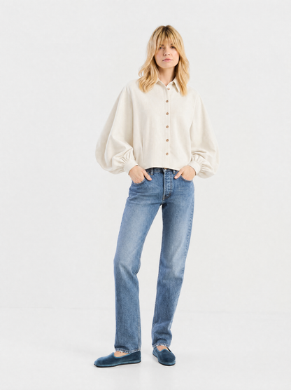 Eine Frau, die eine Bluse „Antonia Short Flanell Offwhite“ mit Ballonärmeln, eine blaue Jeans mit geradem Bein und blaue Samtballerinas trägt, steht vor einem hellgrauen Hintergrund.