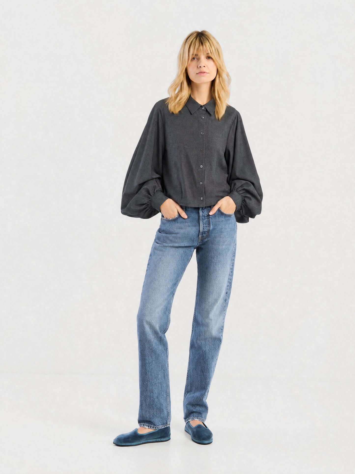 Eine Frau mit blonden Haaren trägt die Bluse „Antonia Short Flanell Anthrazit“ mit voluminösen Ärmeln, dazu eine blaue Jeans. Sie hat die Hände in den Taschen und rundet den Look mit blauen Samtpantoffeln ab.