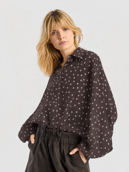 Eine Frau mit blonden Haaren trägt die Bluse „Antonia Polka Dots Brown Ecru“, eine locker sitzende, elegante Bluse aus Baumwollvoile, die in Italien hergestellt wurde. Die Bluse zeichnet sich durch dezente, helle Polka-Dots und weite Ärmel aus und wird zu einer Hose mit hoher Taille kombiniert. Vor einem schlichten Hintergrund steht sie ganz entspannt mit den Händen in den Taschen da.