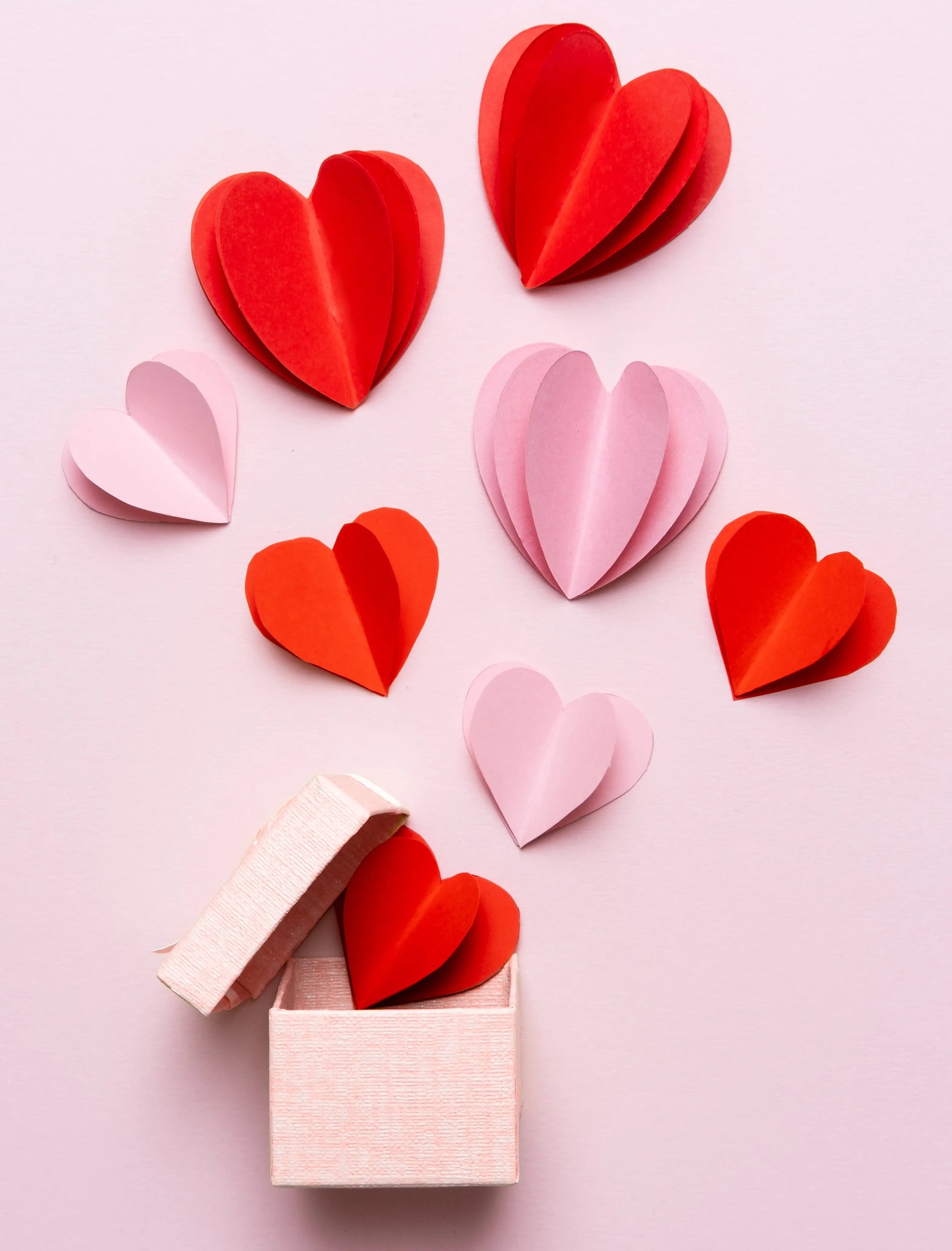 Eine rosafarbene Geschenkschachtel mit abgenommenem Deckel und einem roten Papierherz im Inneren sitzt zwischen roten und rosafarbenen Herzen auf einem hellrosa Hintergrund - ein bezaubernder Geschenkgutschein für einen besonderen Menschen.
