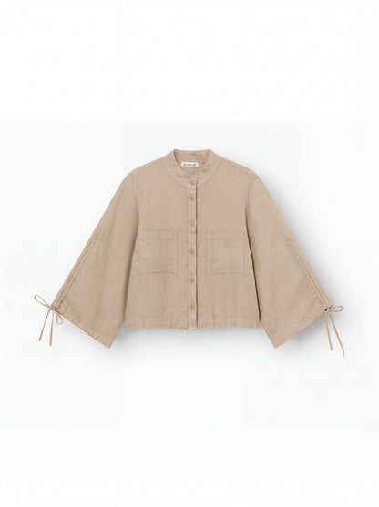 Die Bluse Mary Linen Beige ist eine beige Leinenbluse mit Knopfleiste, weitem Schnitt, verstellbaren Ärmeln samt Schleifendetails, Rundkragen und zwei aufgesetzten Fronttaschen.